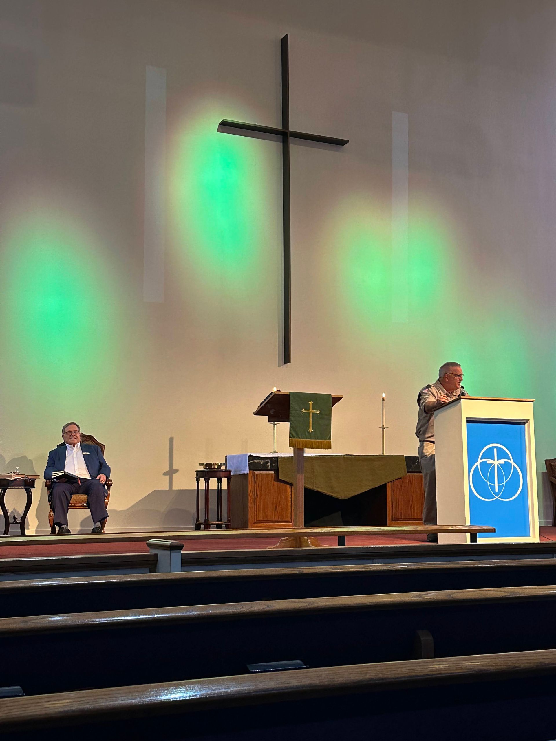 Church interior; man speaking at podium, another seated; cross on wall, green spotlights.