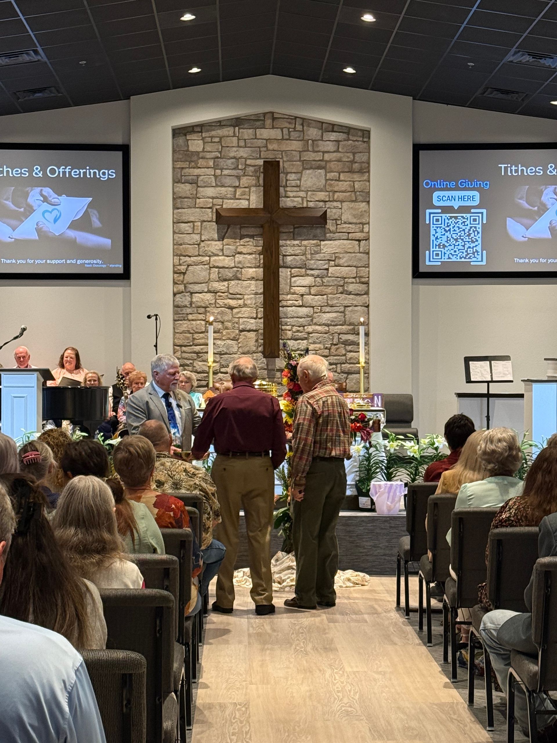 Church service with people seated, a speaker, and a choir. Poinsettias and a cross painting are visible.