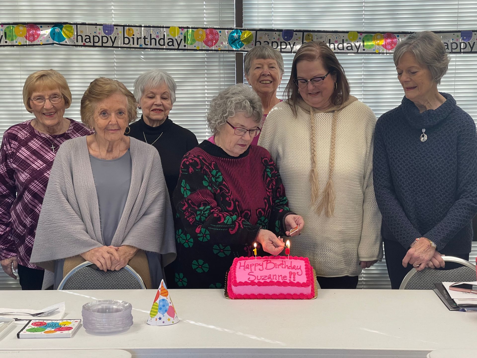 Group of women at a birthday party, lighting candles on a pink cake.