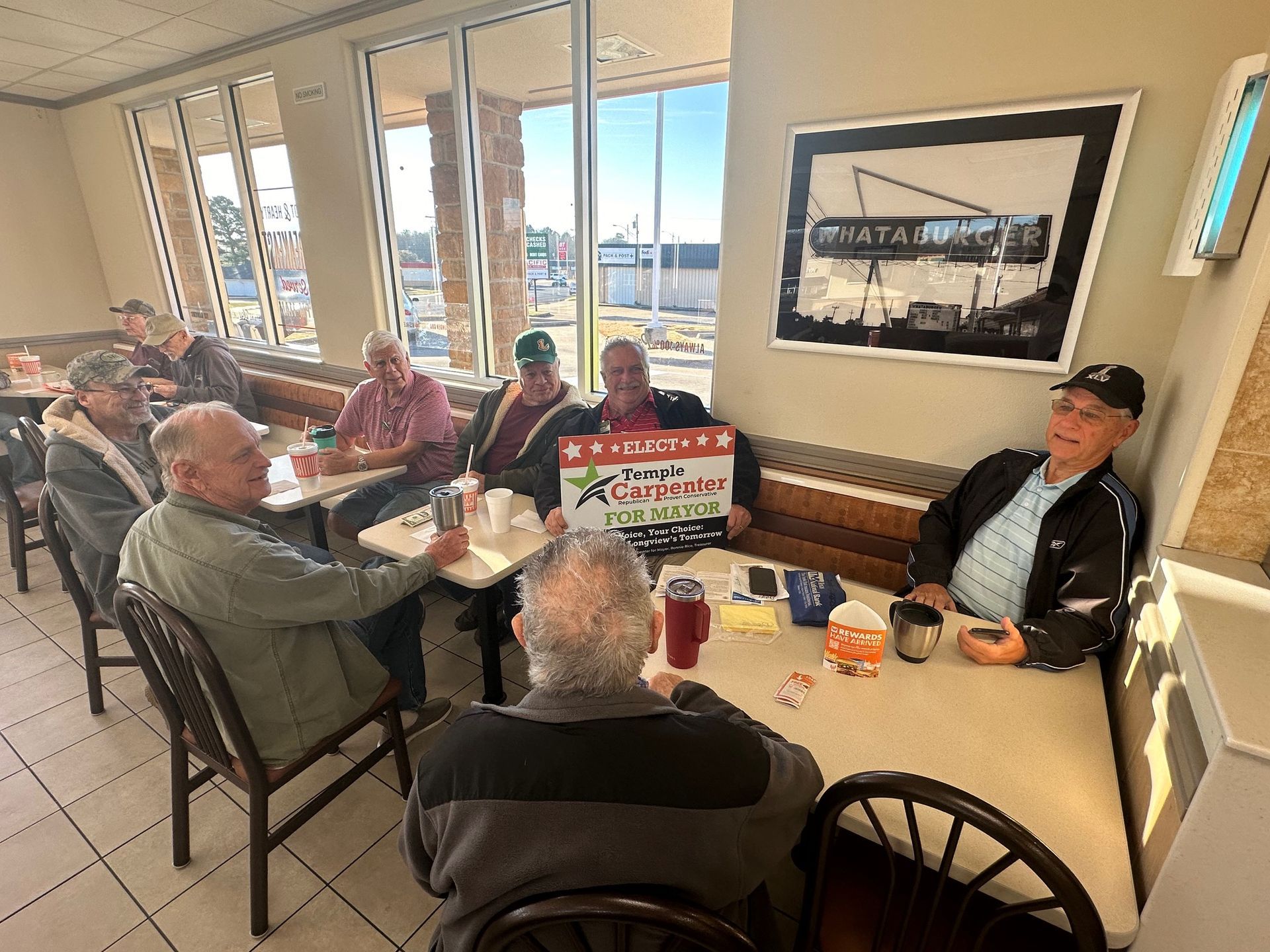 Group of people at a restaurant table holding a sign. Sunlight streams in.
