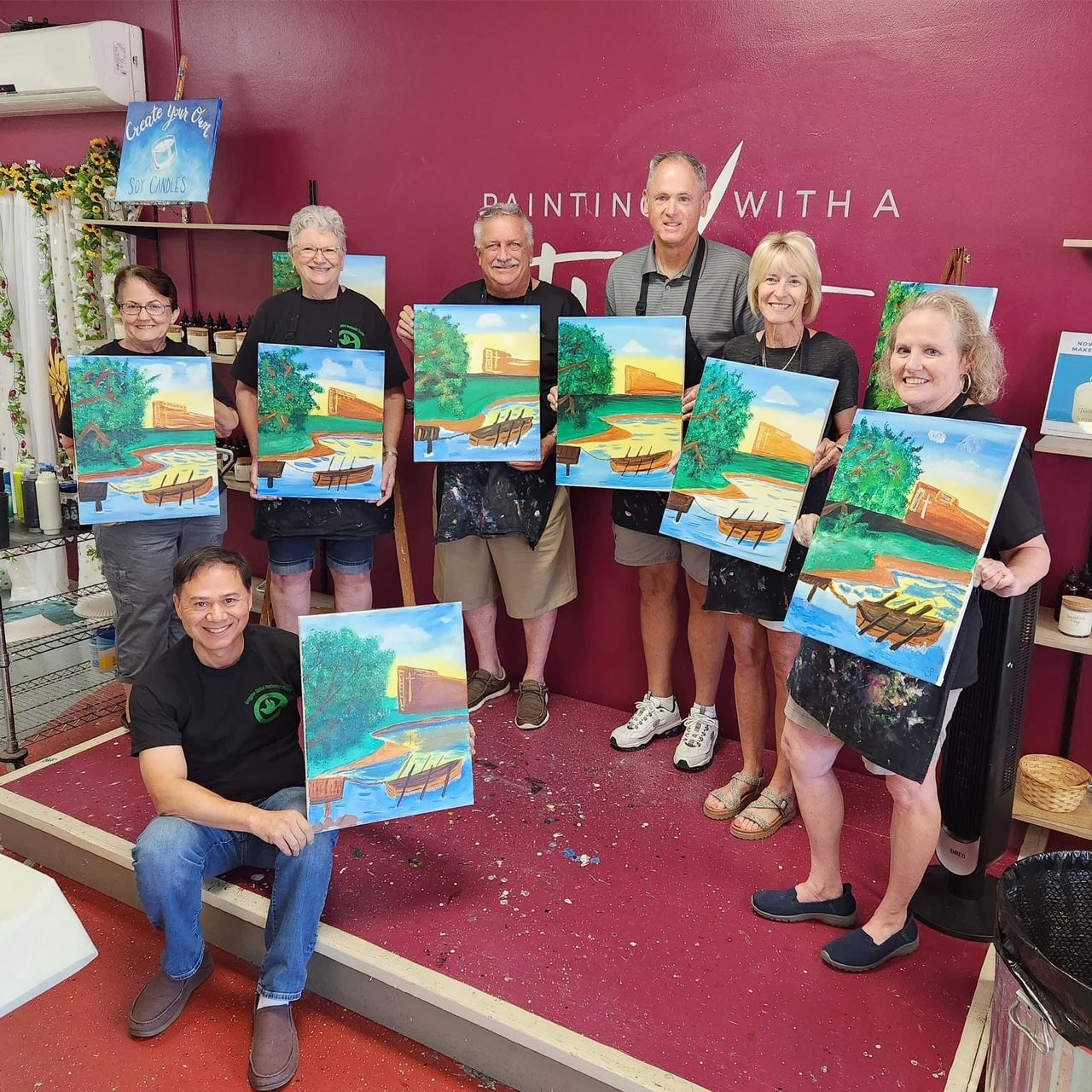 Group of people in a painting class, holding up finished landscape paintings. Inside a studio, with art supplies visible.