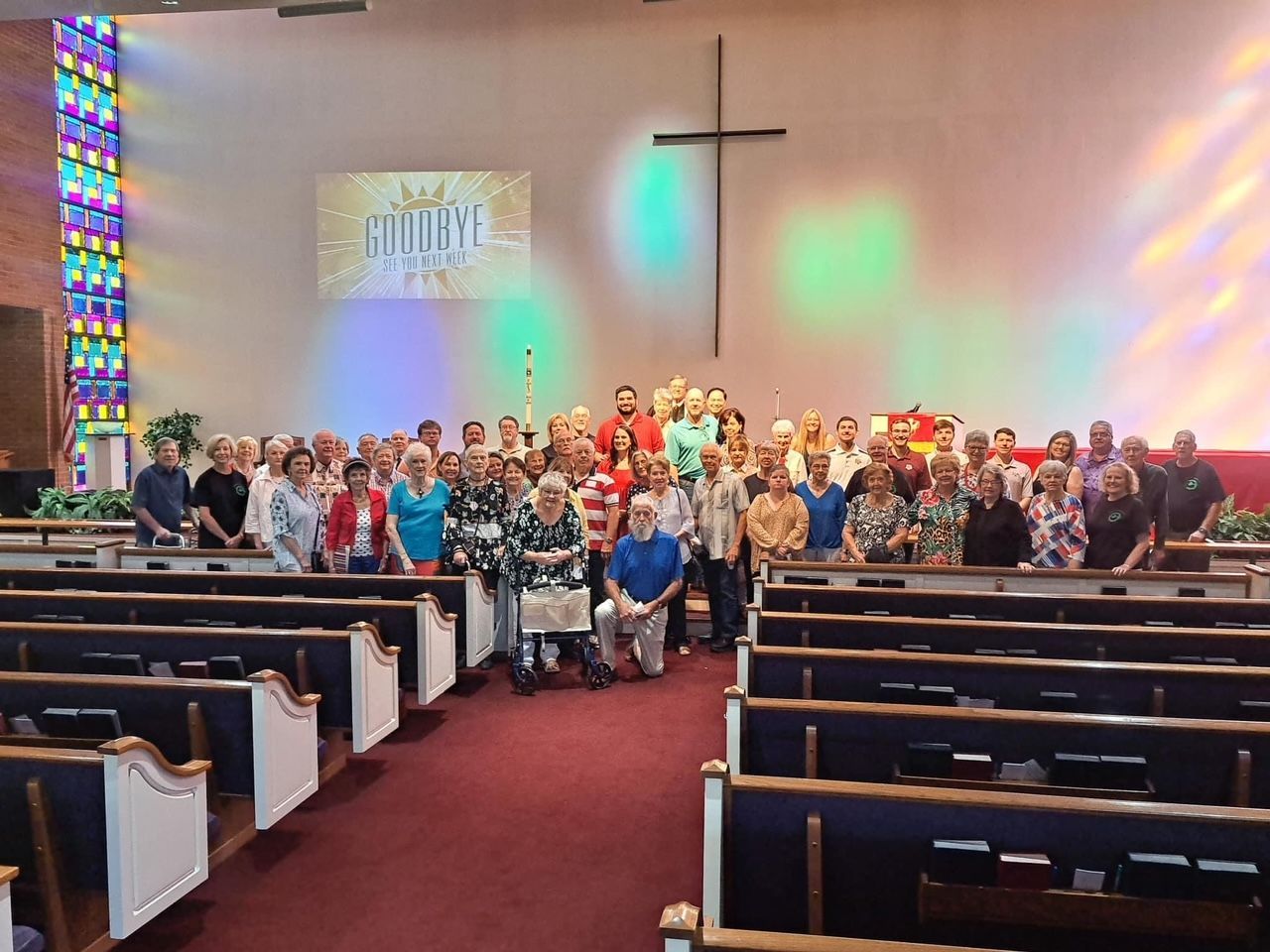 Large group of people standing in a church. A cross hangs on the wall, and colored light shines.