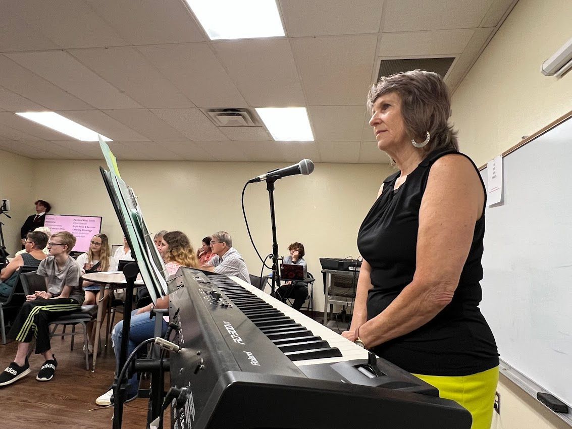 Woman at keyboard with microphone, facing audience. A classroom setting with other people present.