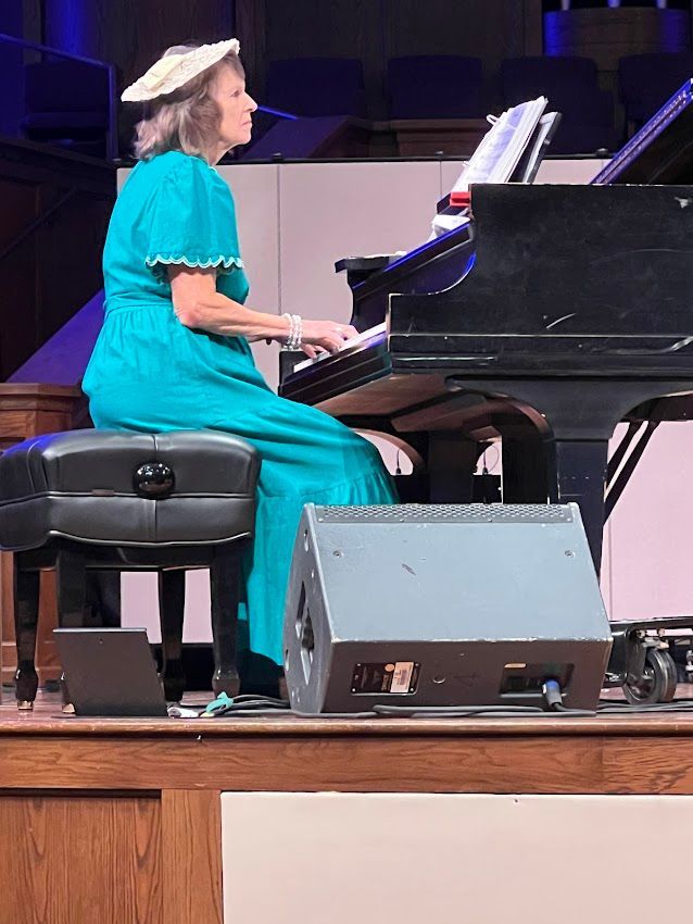 Woman in teal dress plays piano on stage, wearing a hat.