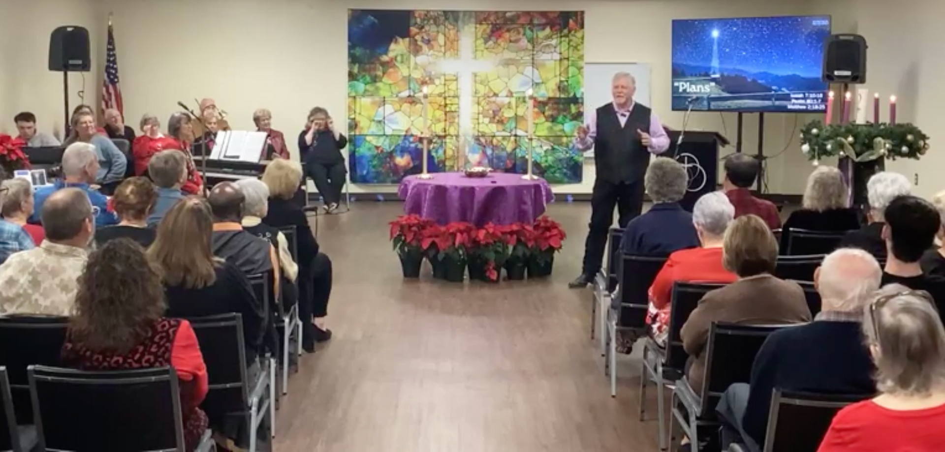 Church service with people seated, a speaker, and a choir. Poinsettias and a cross painting are visible.