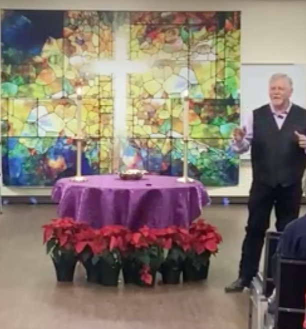 Church service with people seated, a speaker, and a choir. Poinsettias and a cross painting are visible.