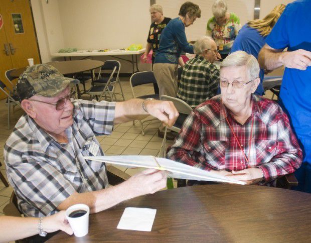 Two men seated, one in camo hat, holding a paper airplane. Others gather in a room, some at a table.
