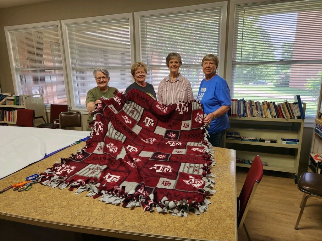 Four women holding a maroon and gray Texas A&M blanket at a table in a room with windows.