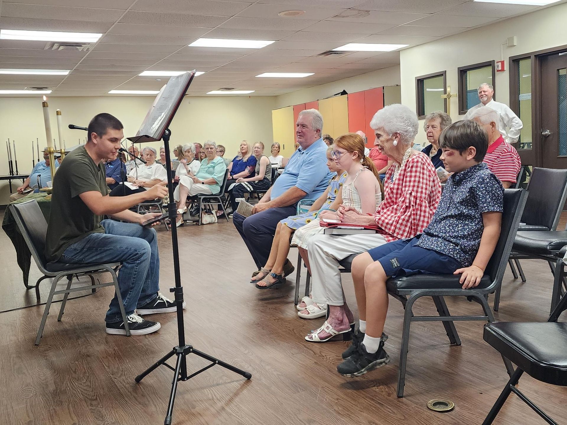 A young man performs for an audience. He sits, gesturing. People of varying ages watch from chairs in a room.