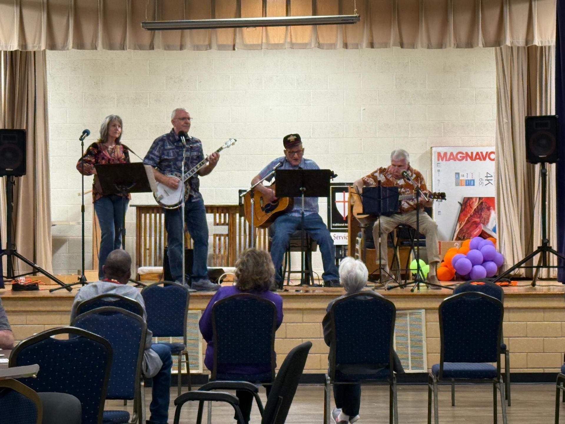 Band playing on stage for seated audience. Musicians with banjo, guitar, accordion. Stage has sound equipment and MAGNAVOX banner.