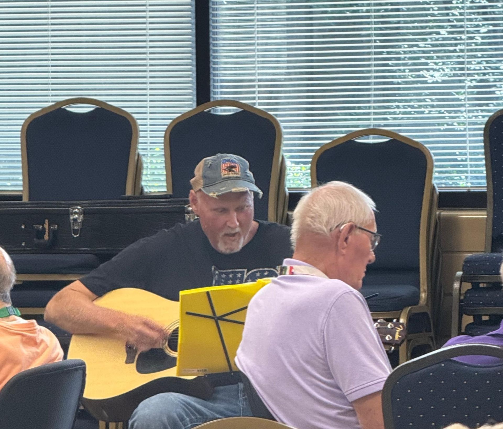 Man plays guitar, sings. Seated in a room with others. Yellow music stand, blue chairs, blinds.