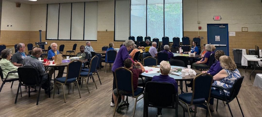 People seated at tables in a community hall. Some are interacting, others appear to be working or playing a game.