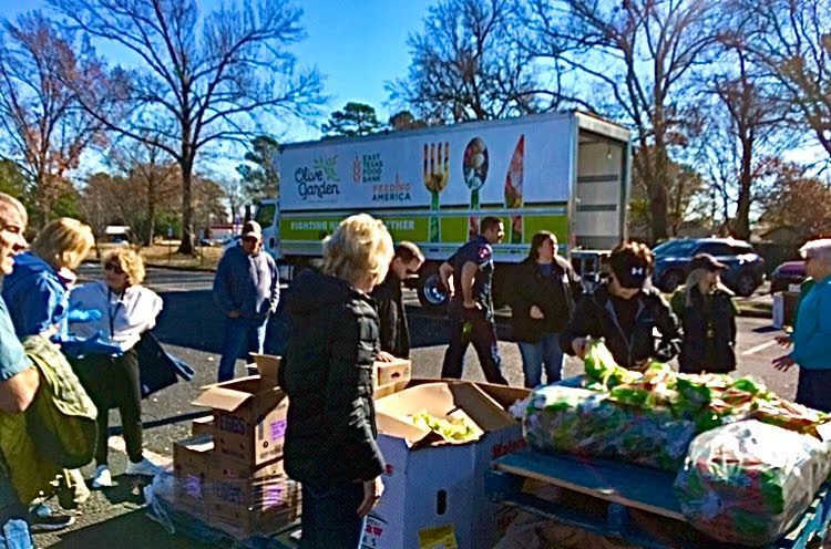 People loading boxes of produce from an Olive Garden truck in a parking lot on a sunny day.