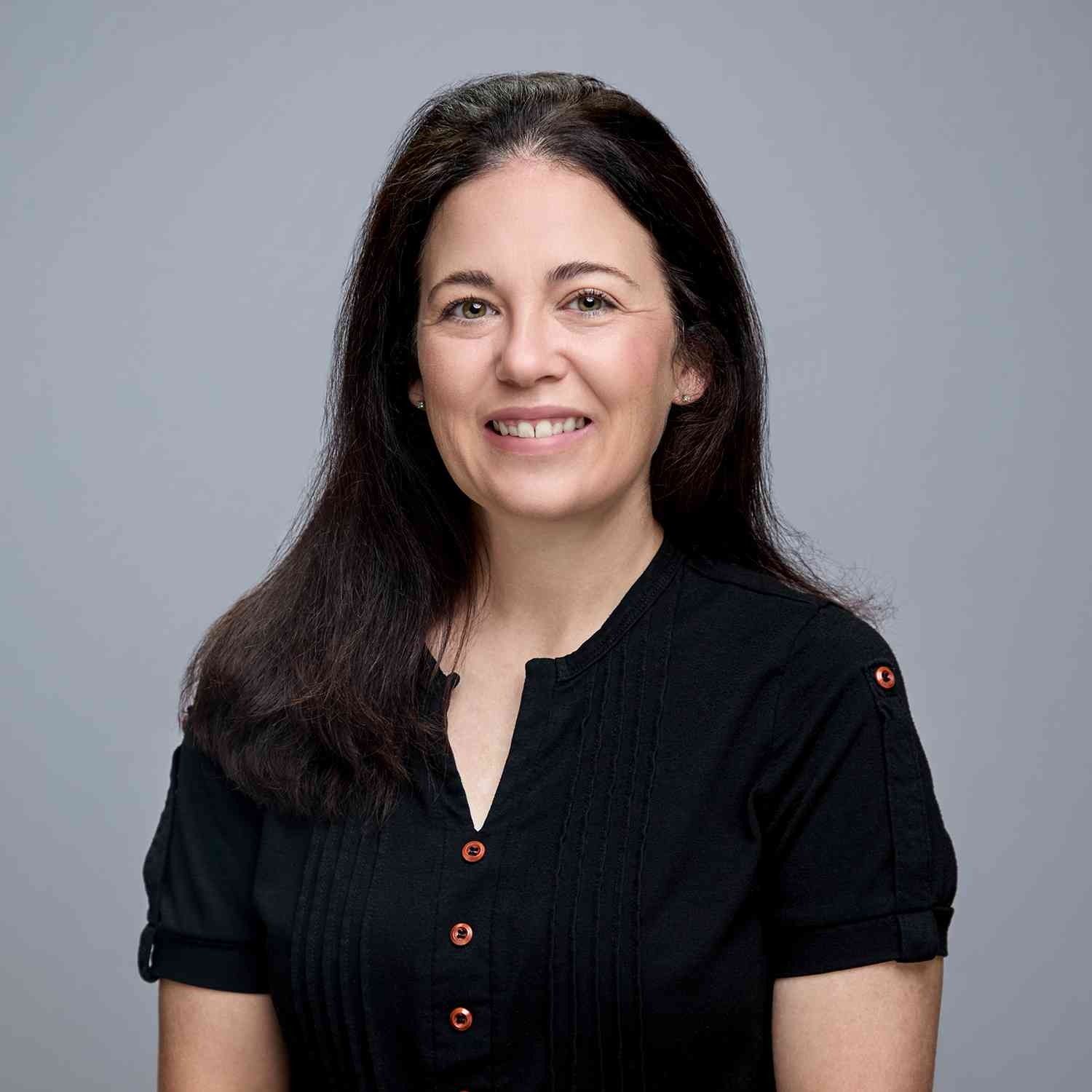 Woman with long, dark hair smiles. She wears a black shirt against a gray backdrop.