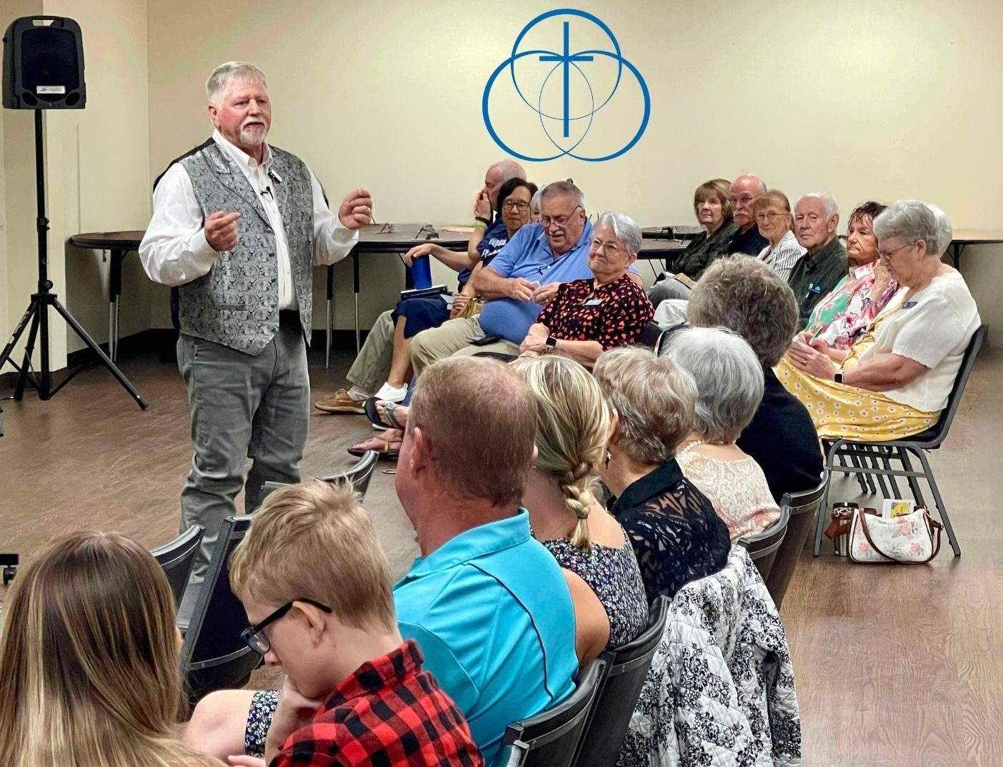 Man speaking to an audience seated in a room, with a logo on the wall.