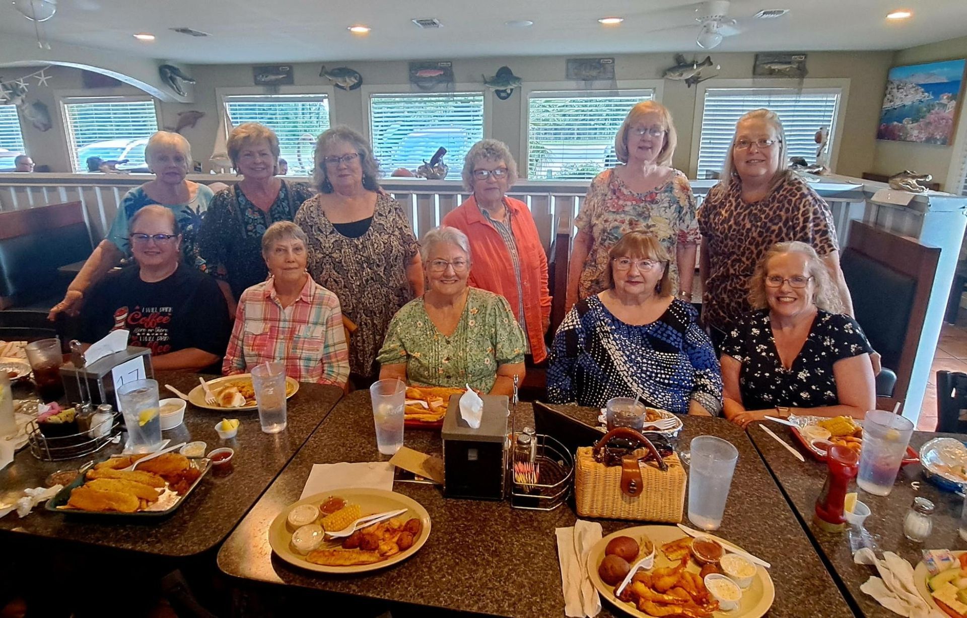 Group of women at a restaurant table with plates of food. Windows in background, bright setting.