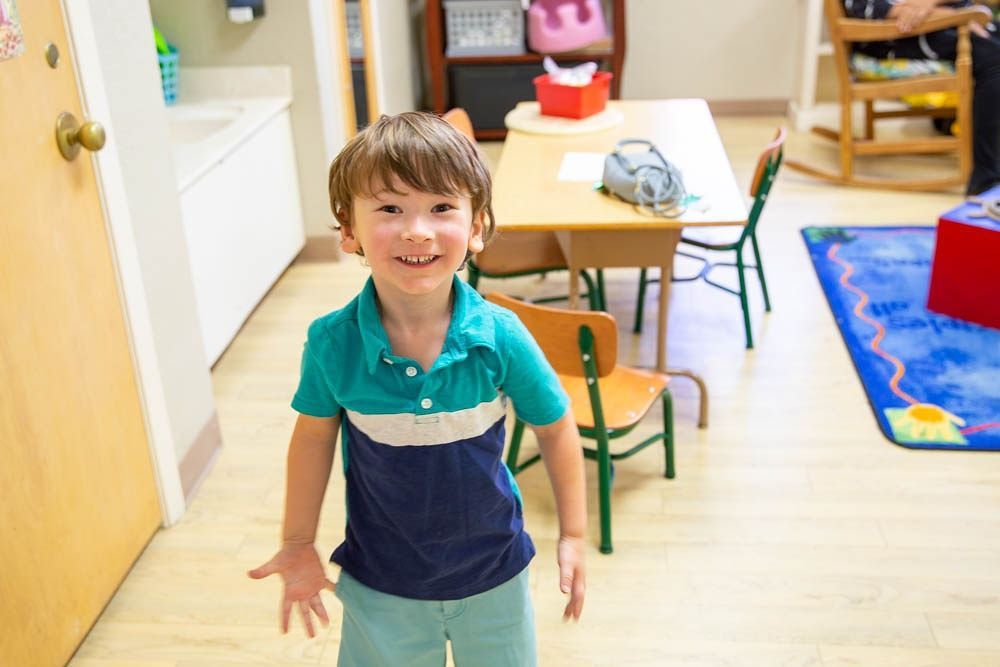 Smiling child in a blue and white shirt stands in a brightly lit room with a table and chairs in the background.