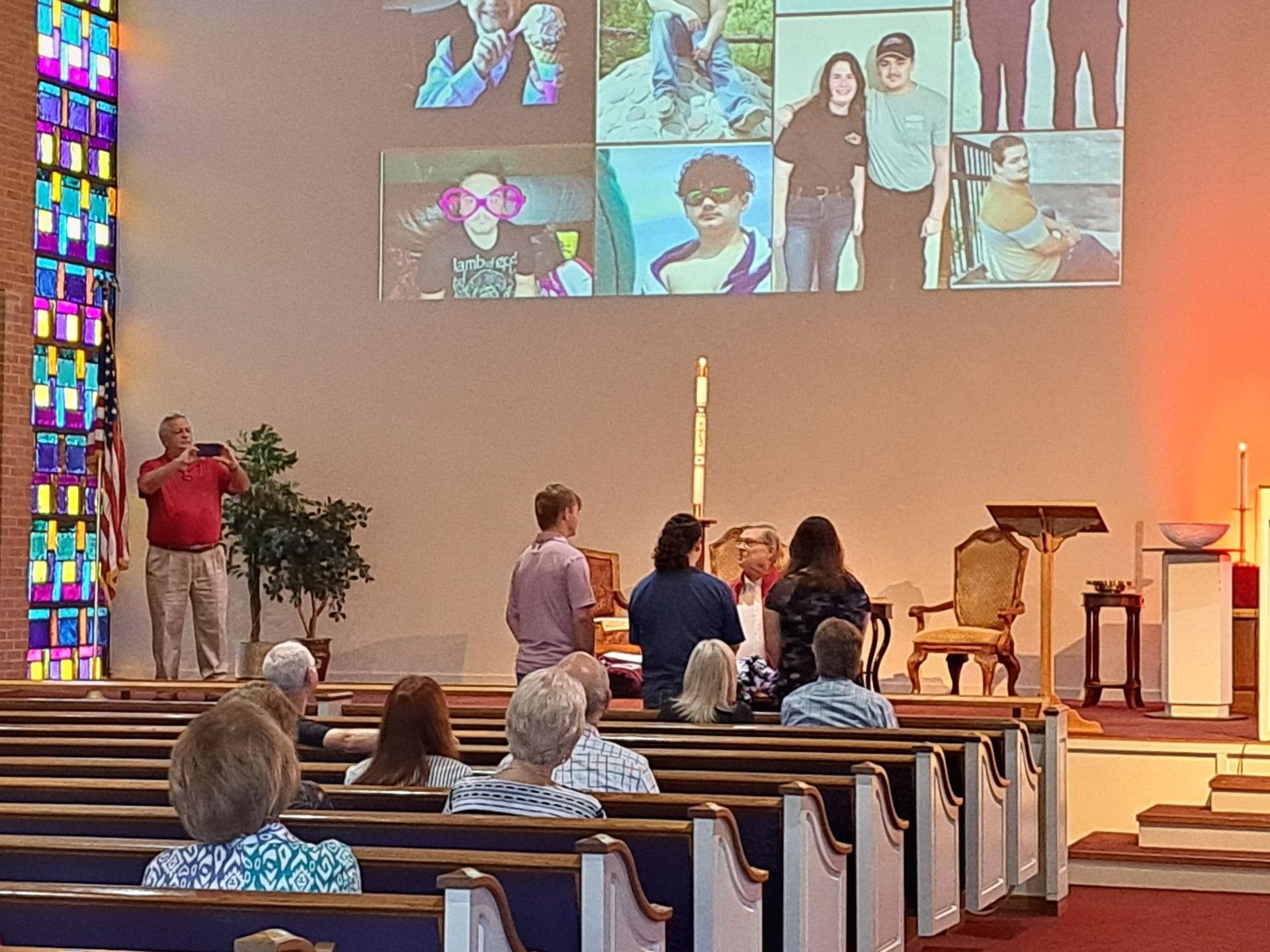 A church service in progress. People seated in pews face a stage with a projection screen.