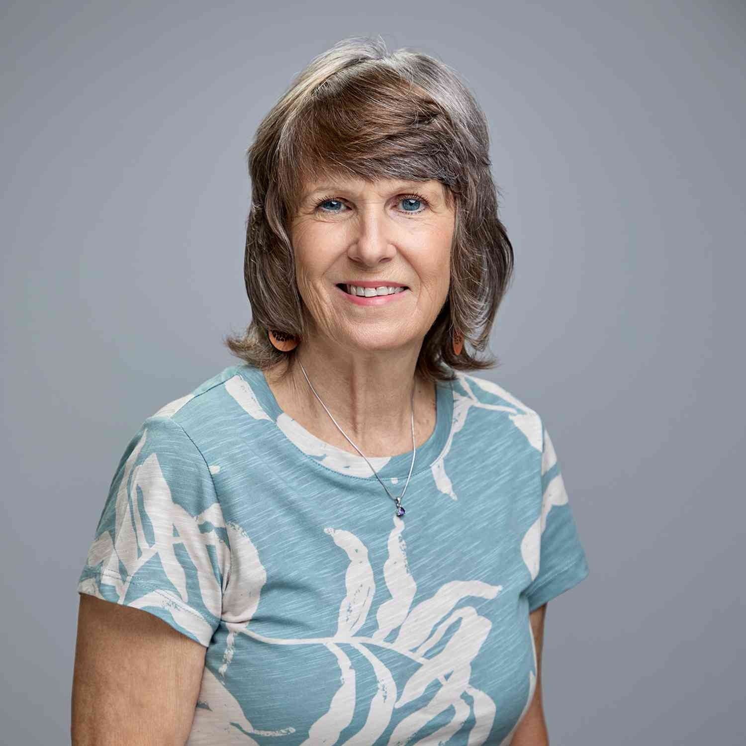 Woman smiling, wearing a blue patterned top, against a gray backdrop.