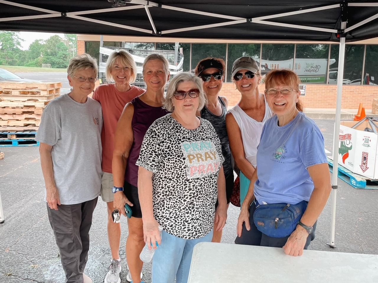 Group of seven people smiling under a black canopy. They stand in front of a building with pallets.