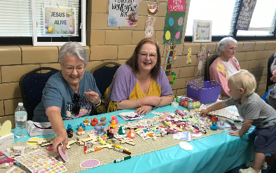 Three women and a child at a craft table; smiling, surrounded by colorful craft supplies and decorations.