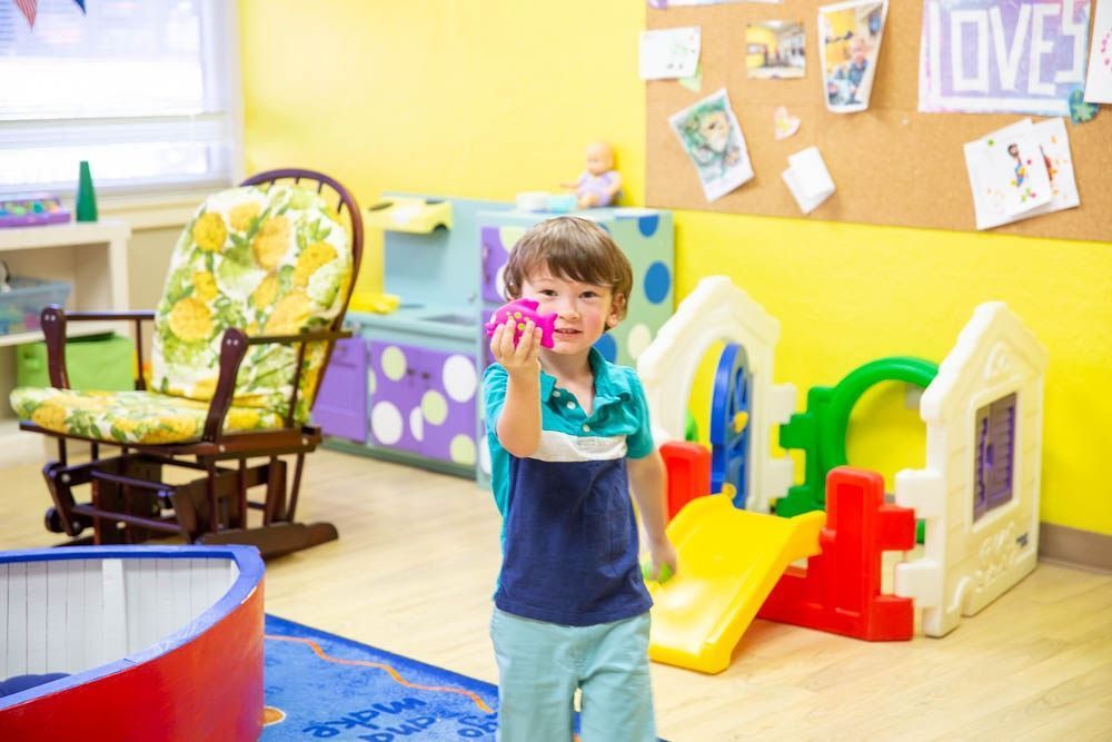 Boy holding a toy, smiles in a brightly colored playroom with playhouse and furniture.