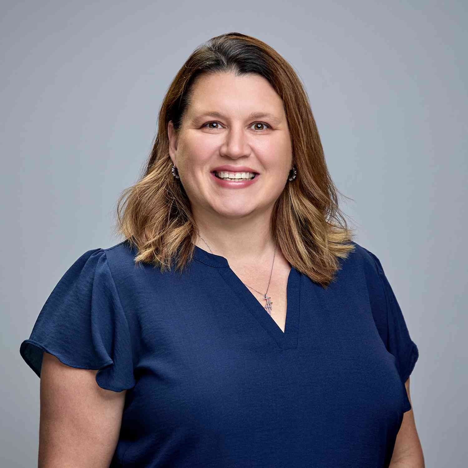 Woman with shoulder-length brown hair, smiling, wearing a navy blouse against a gray backdrop.