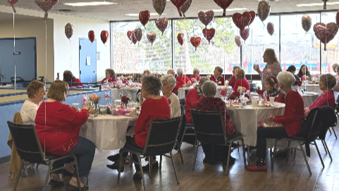 Group of people in red at a decorated Valentine's Day event. Heart balloons hang from the ceiling.