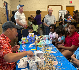 People at a craft table in a room. Someone is making a craft with kids. Others watch.