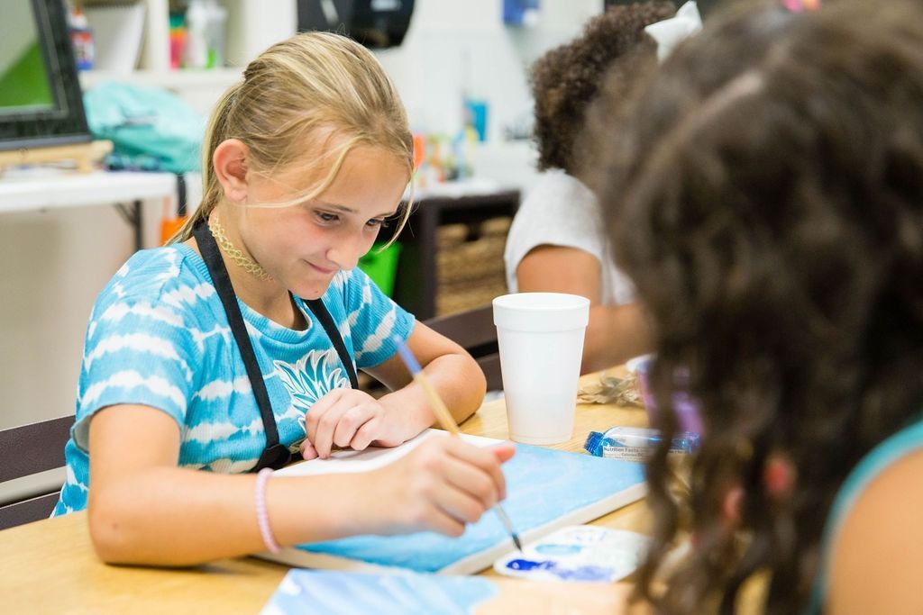 Girl in a blue tie-dye shirt painting at a table, focused expression. Other children are partially visible.