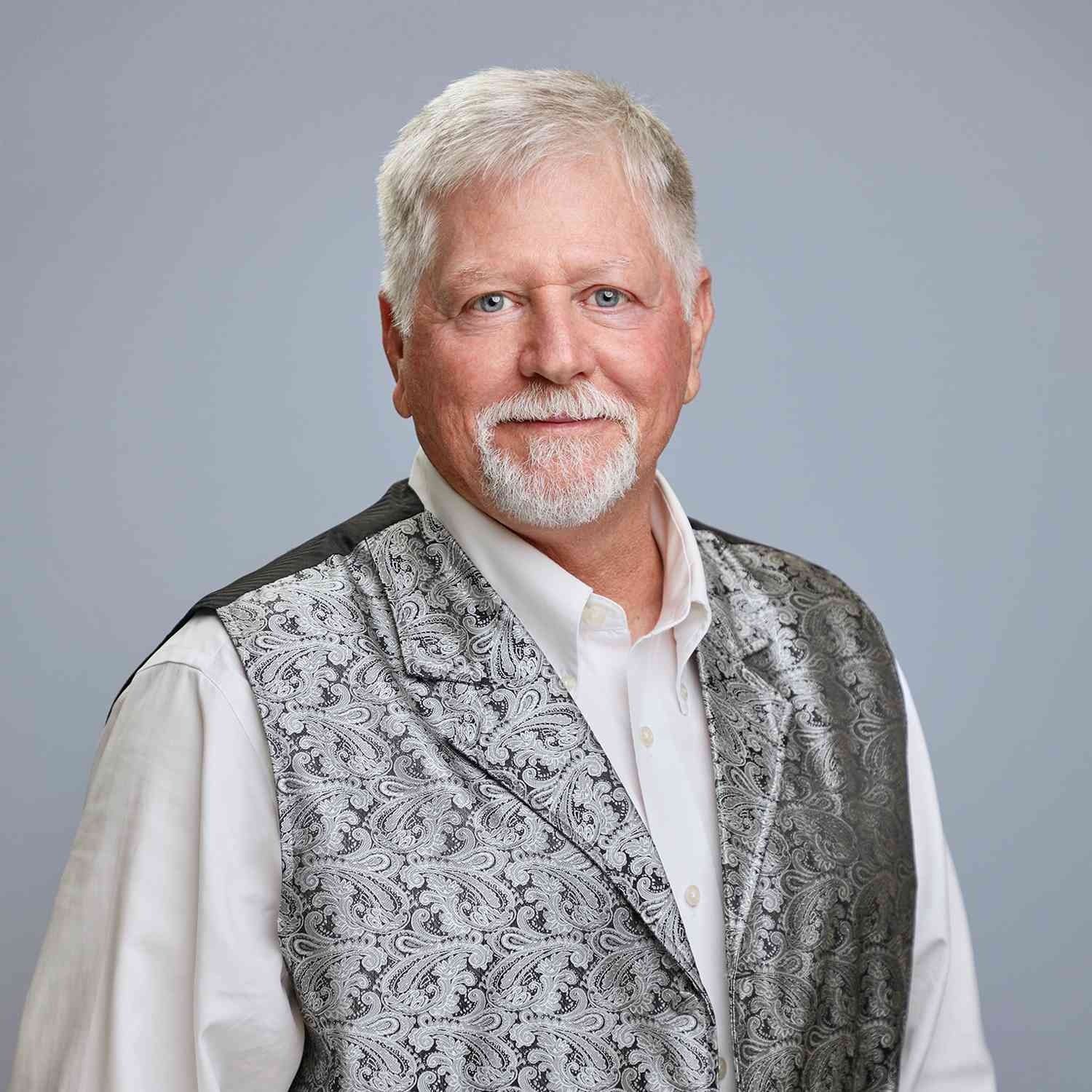 Man with white hair and goatee, wearing a patterned vest and white shirt, smiling.