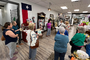 People gathered inside a store, listening to a woman. A Texas flag hangs on the wall.