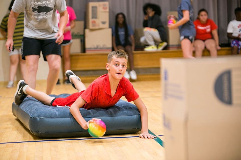 Boy in red shirt on inflatable, holding ball, playing game in gym. Others watch, boxes in background.