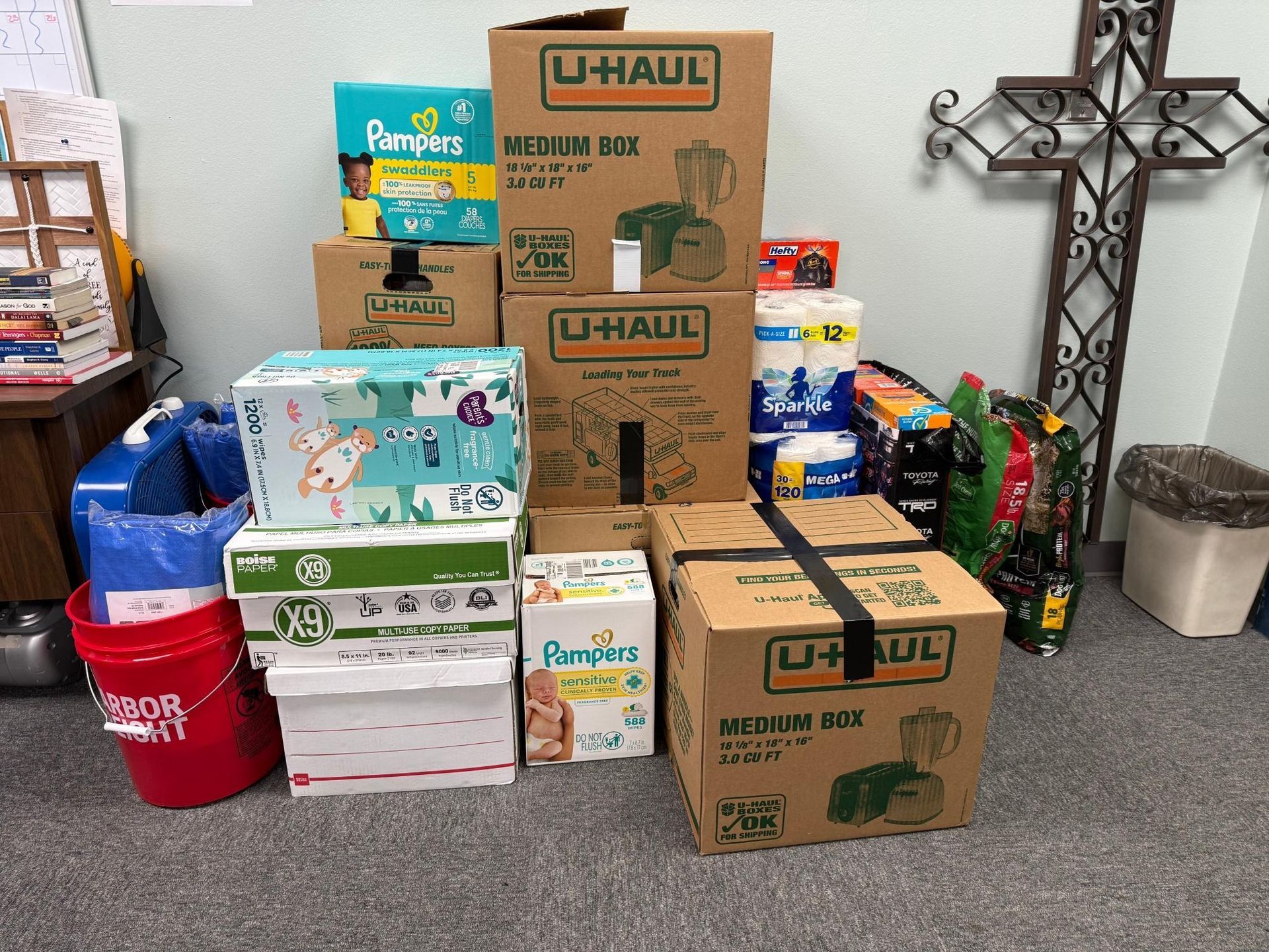 Stack of donation boxes, including U-Haul boxes, diapers, and supplies, near a cross.