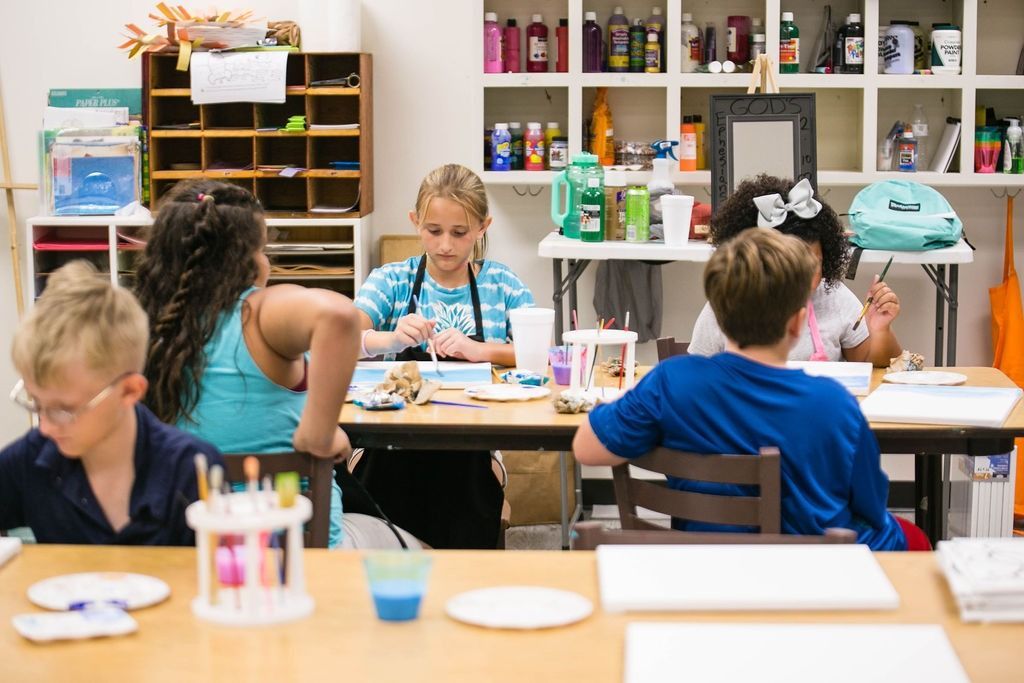 Children painting at a table in an art room. Paint supplies on shelves.