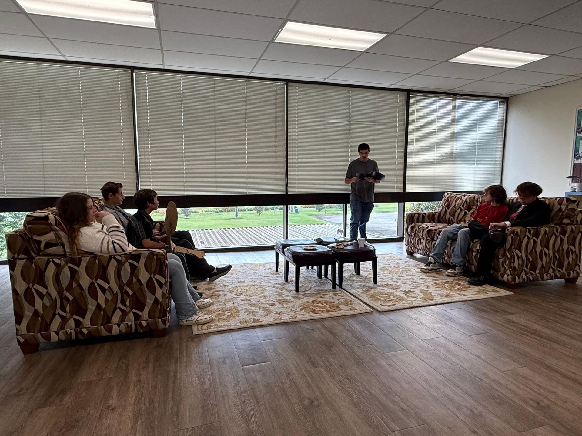 People gathered in a room, with blinds and a window. A person stands reading in front of others seated on couches.
