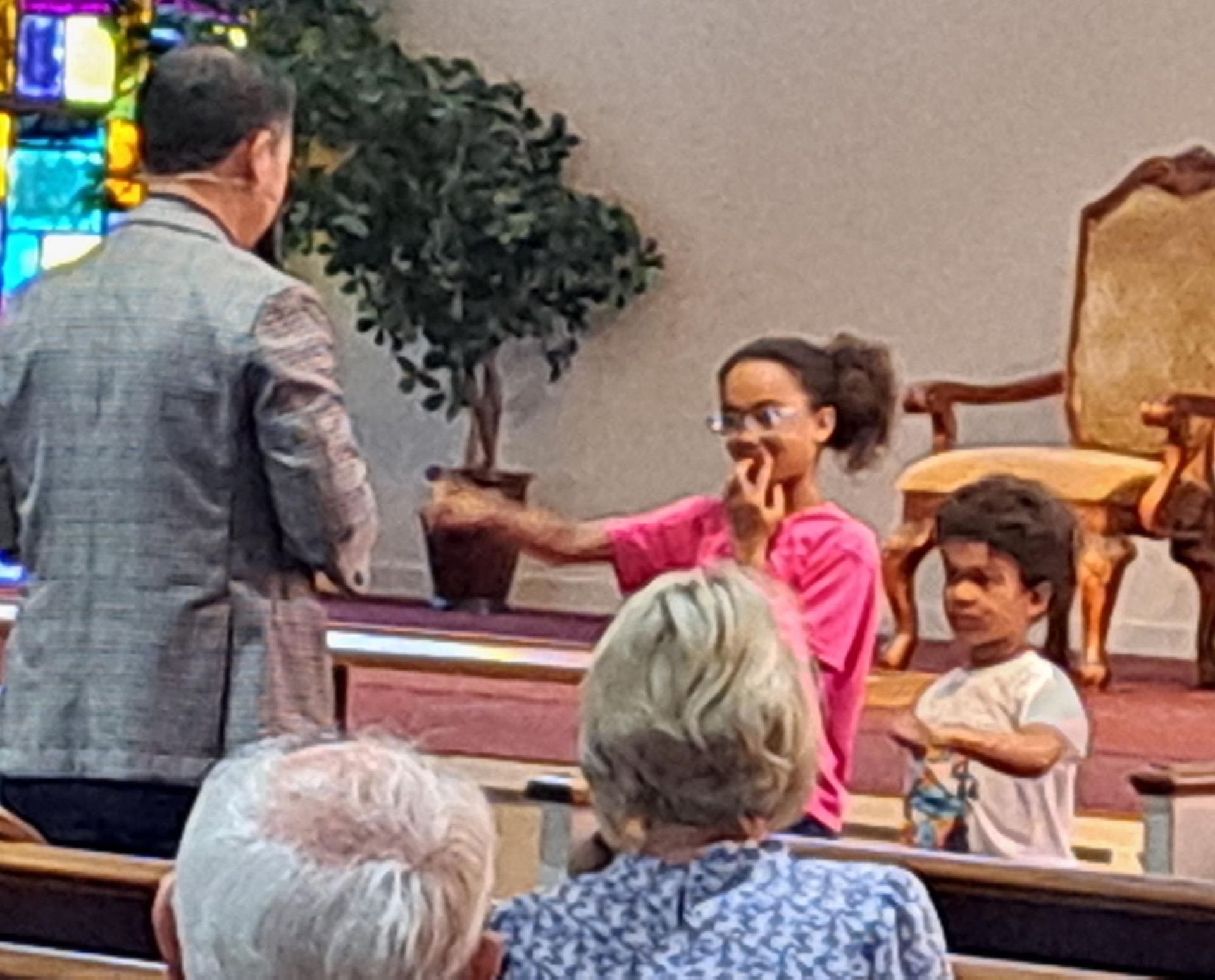 Inside a church, a person in a gray jacket speaks to a child holding an object. Another child stands nearby.