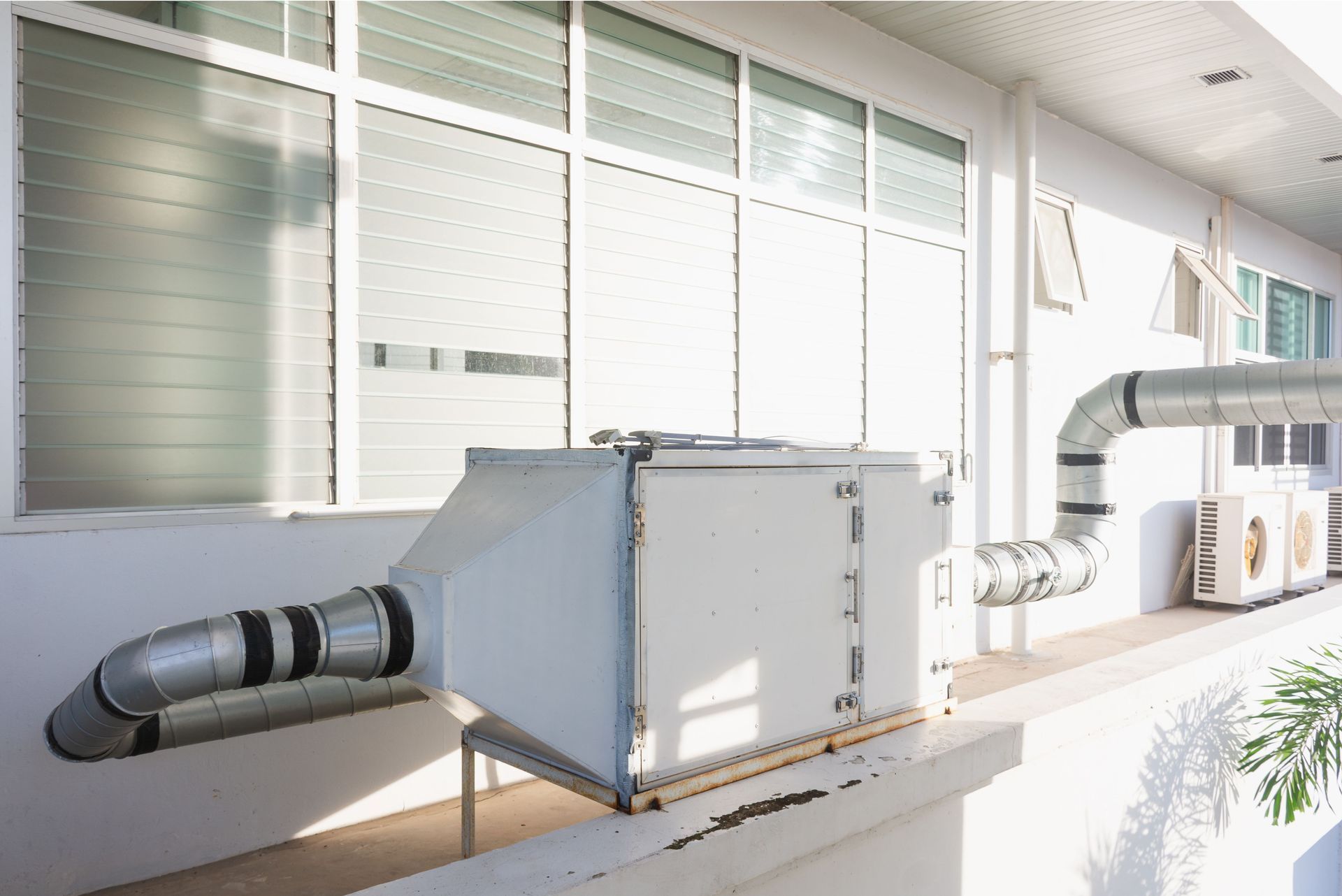 A white box with pipes attached to it is on the side of a building next to a window.