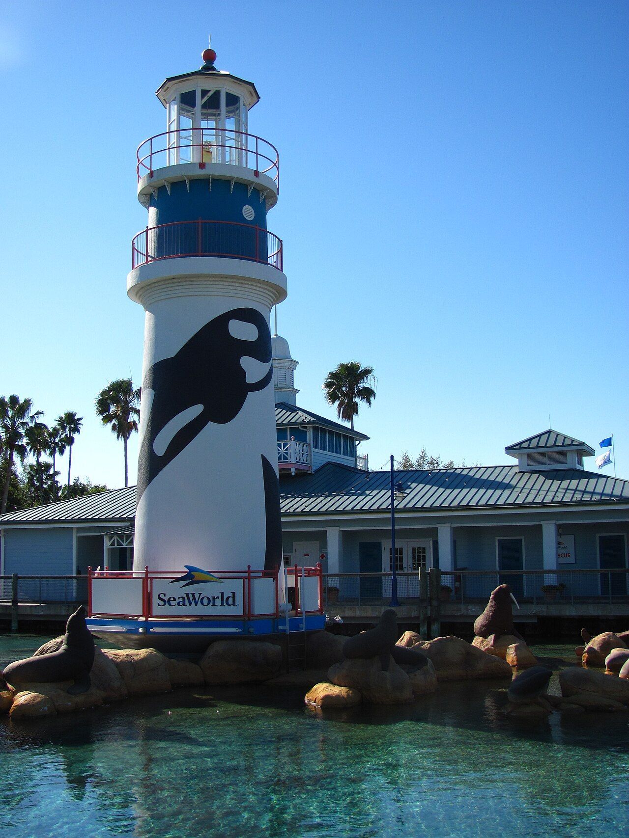 Tall white lighthouse with black dolphin mural beside turquoise water and palm trees under blue sky