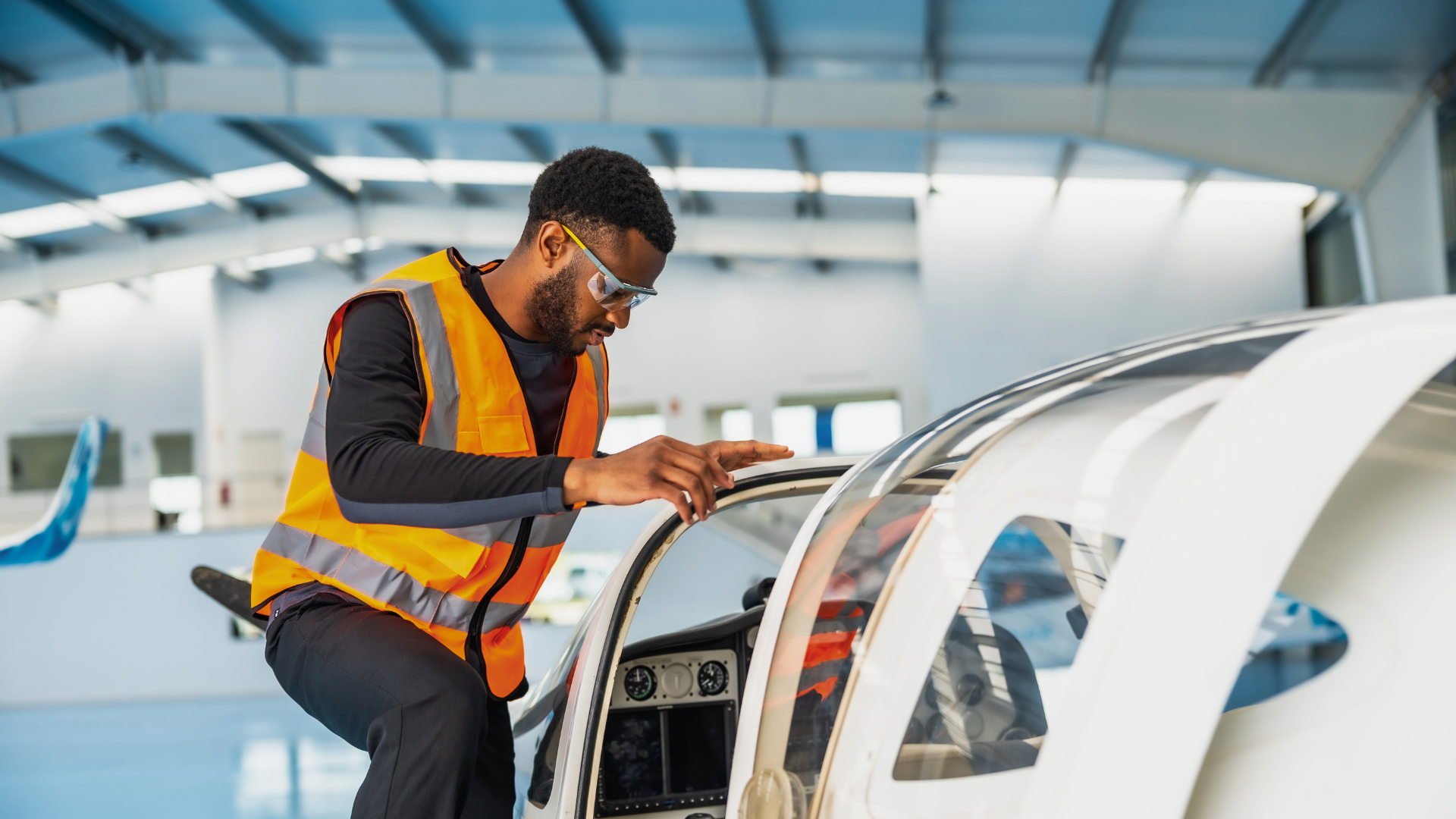A technician in a high-visibility vest inspects the cockpit of a light aircraft inside an airplane hangar.