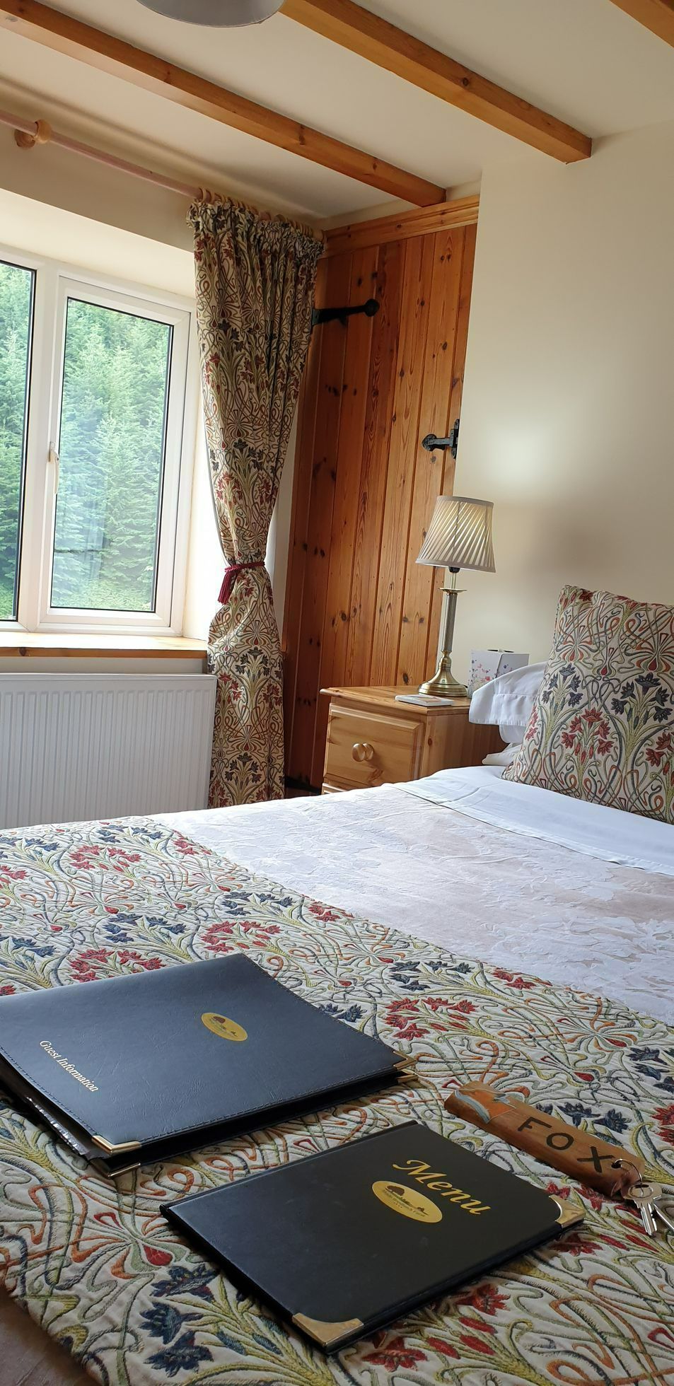 A cozy bedroom with a patterned bedspread, two black books, a window, and wooden beams on the ceiling.