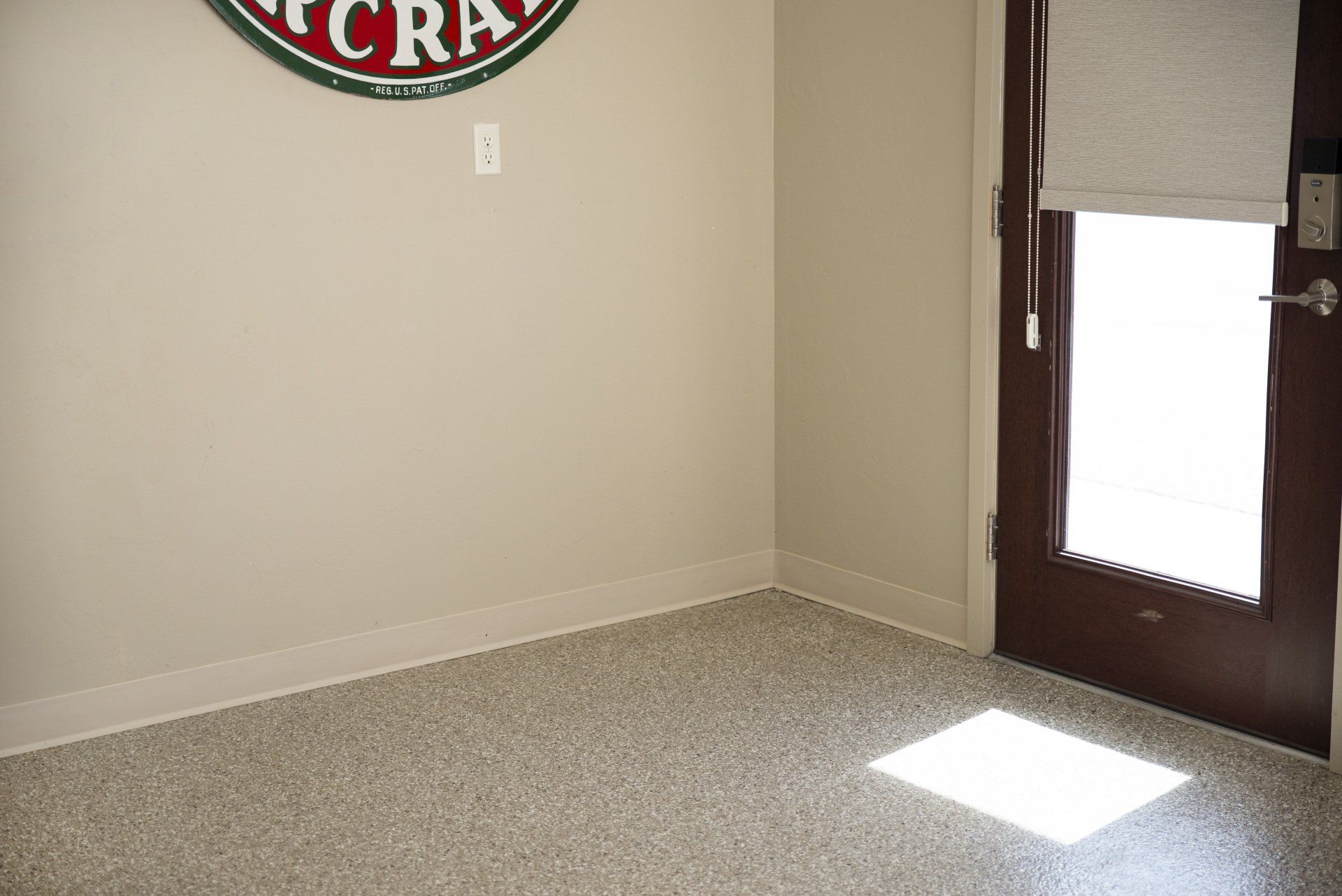 Corner of a room with speckled flooring, beige walls, and a brown door with a window letting in sunlight.
