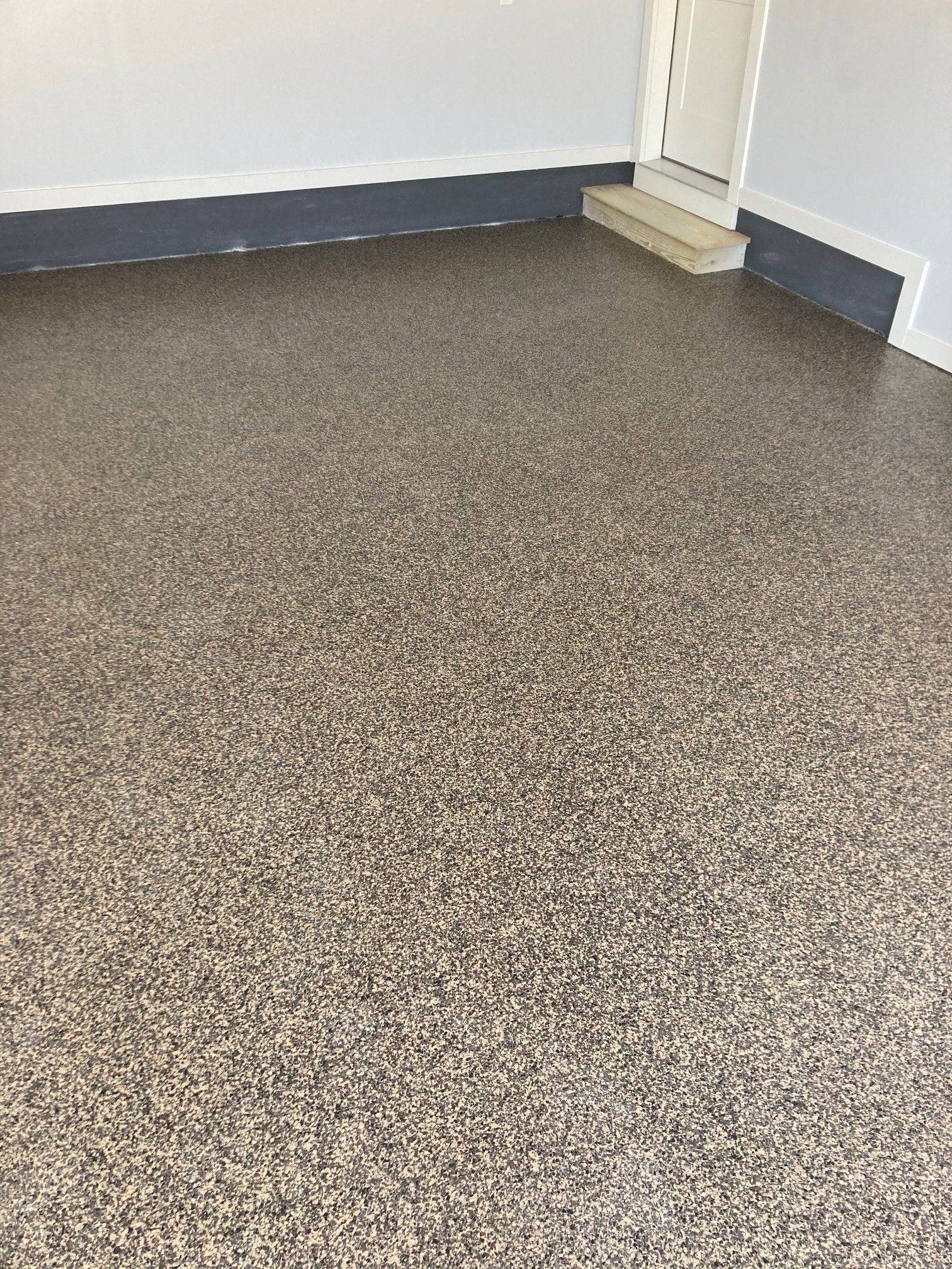 Garage with a speckled, brown epoxy floor. Gray walls and a dark gray baseboard. White door in the background.