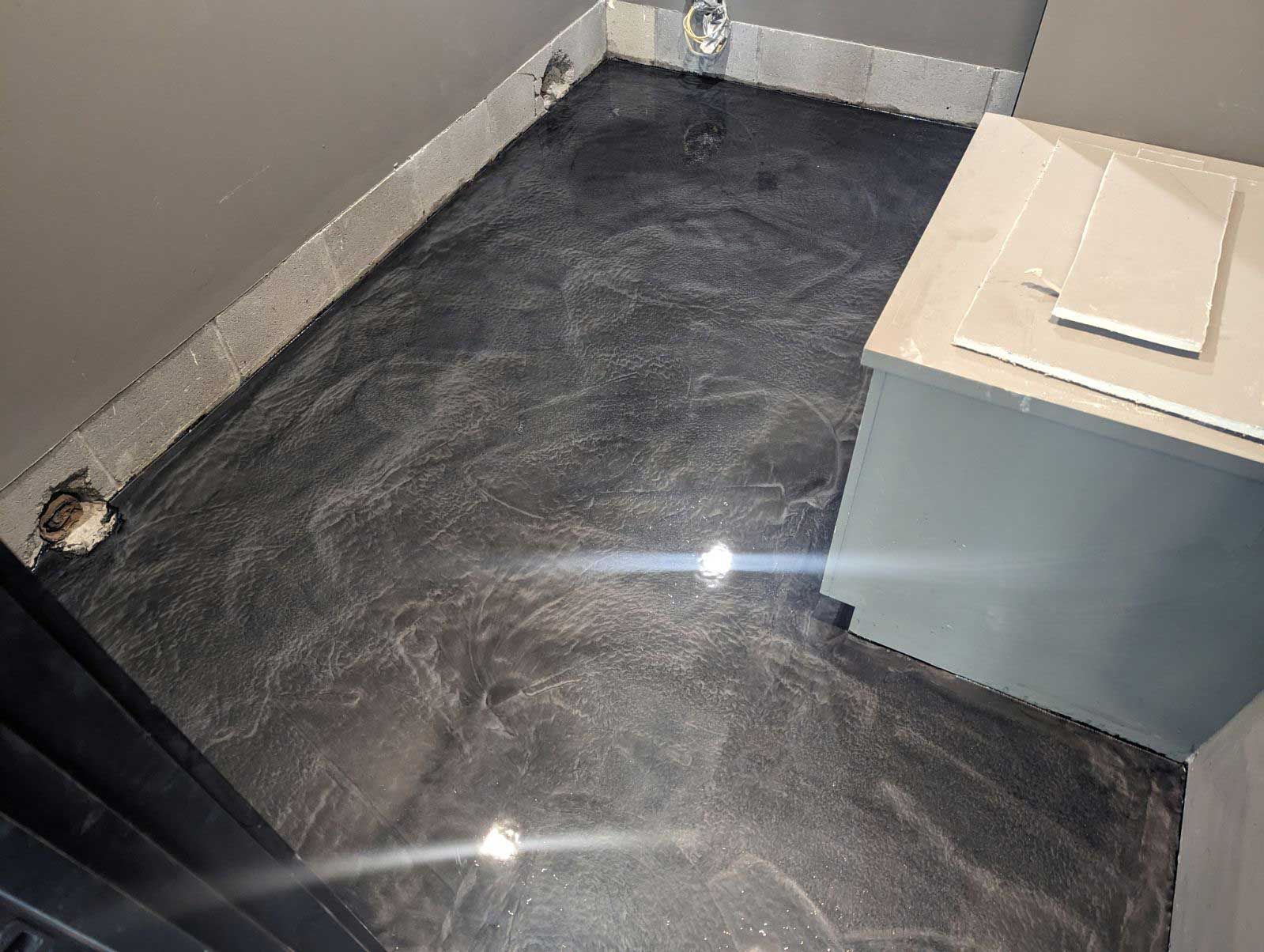 A small bathroom with a dark, shiny, metallic-looking floor. The walls are a lighter gray with a corner cabinet on the right.