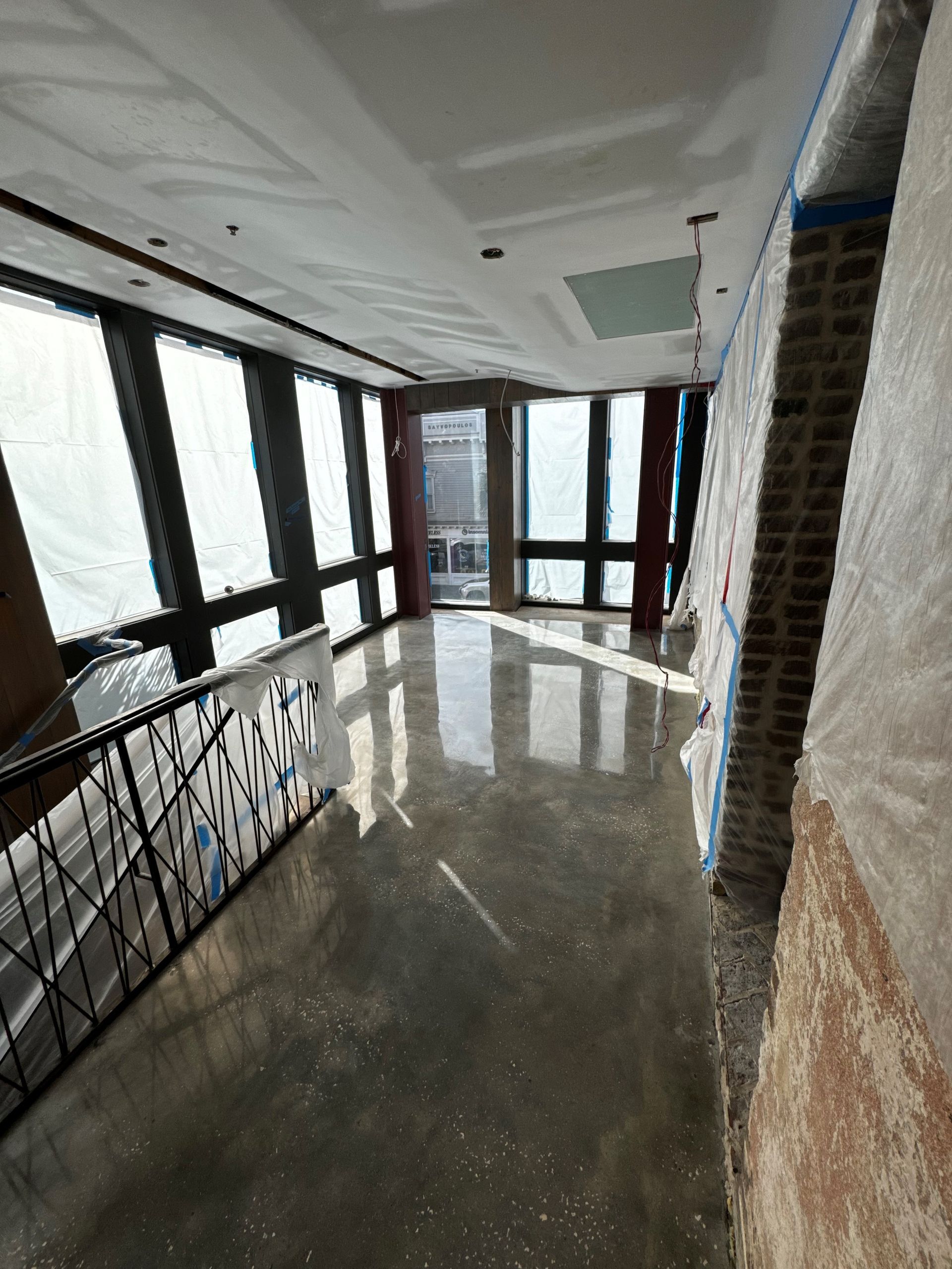 Interior space under construction with large windows, a brick wall, and a concrete floor. 