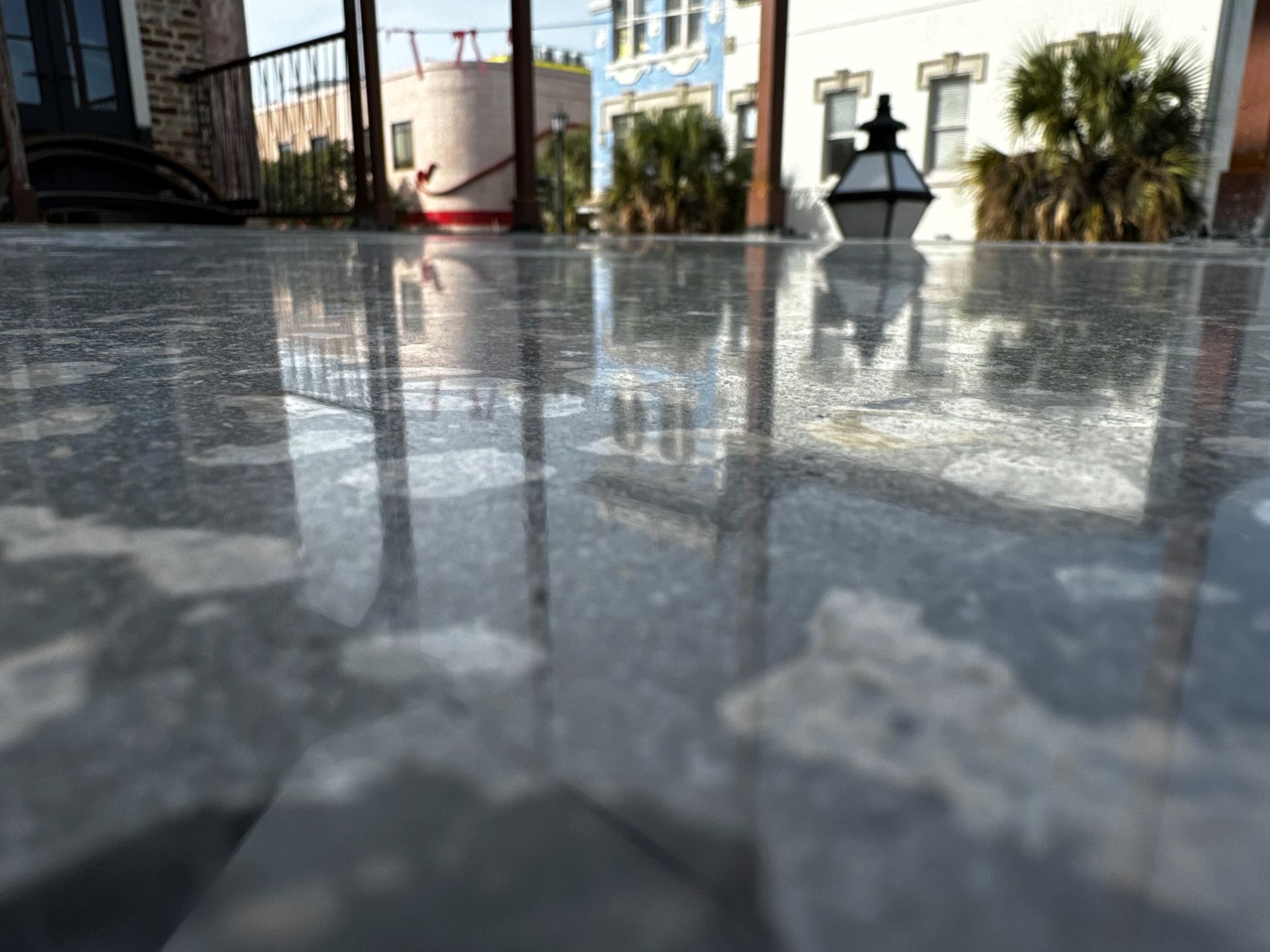 Close-up of a reflective, gray stone surface reflecting nearby buildings, a light post, and palm trees in a city setting.
