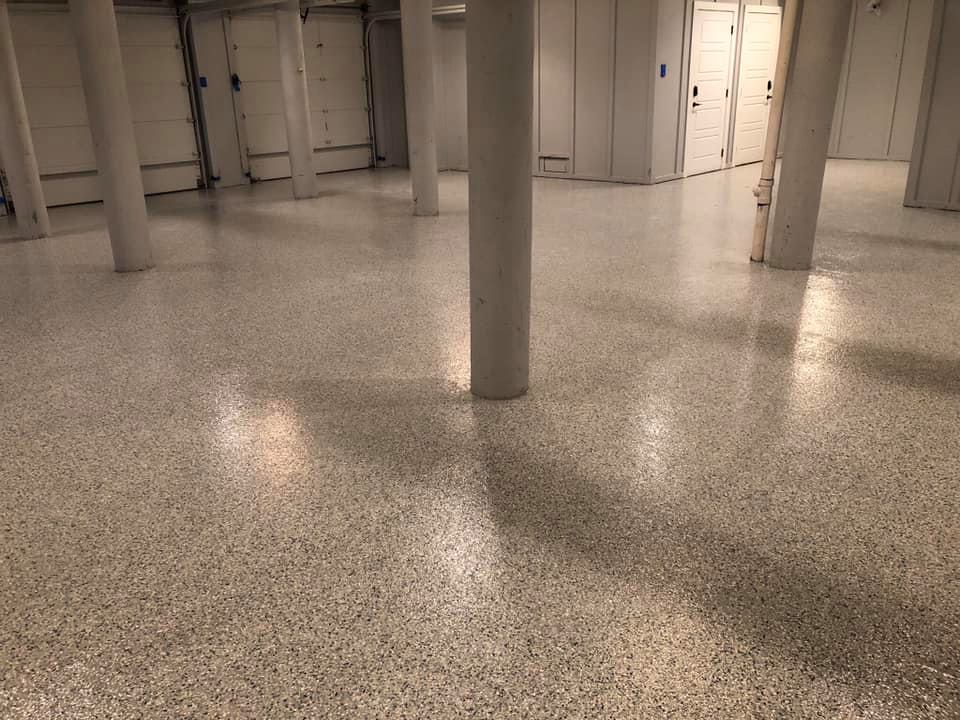 Light-colored, speckled epoxy flooring in a finished basement with white support columns and walls.