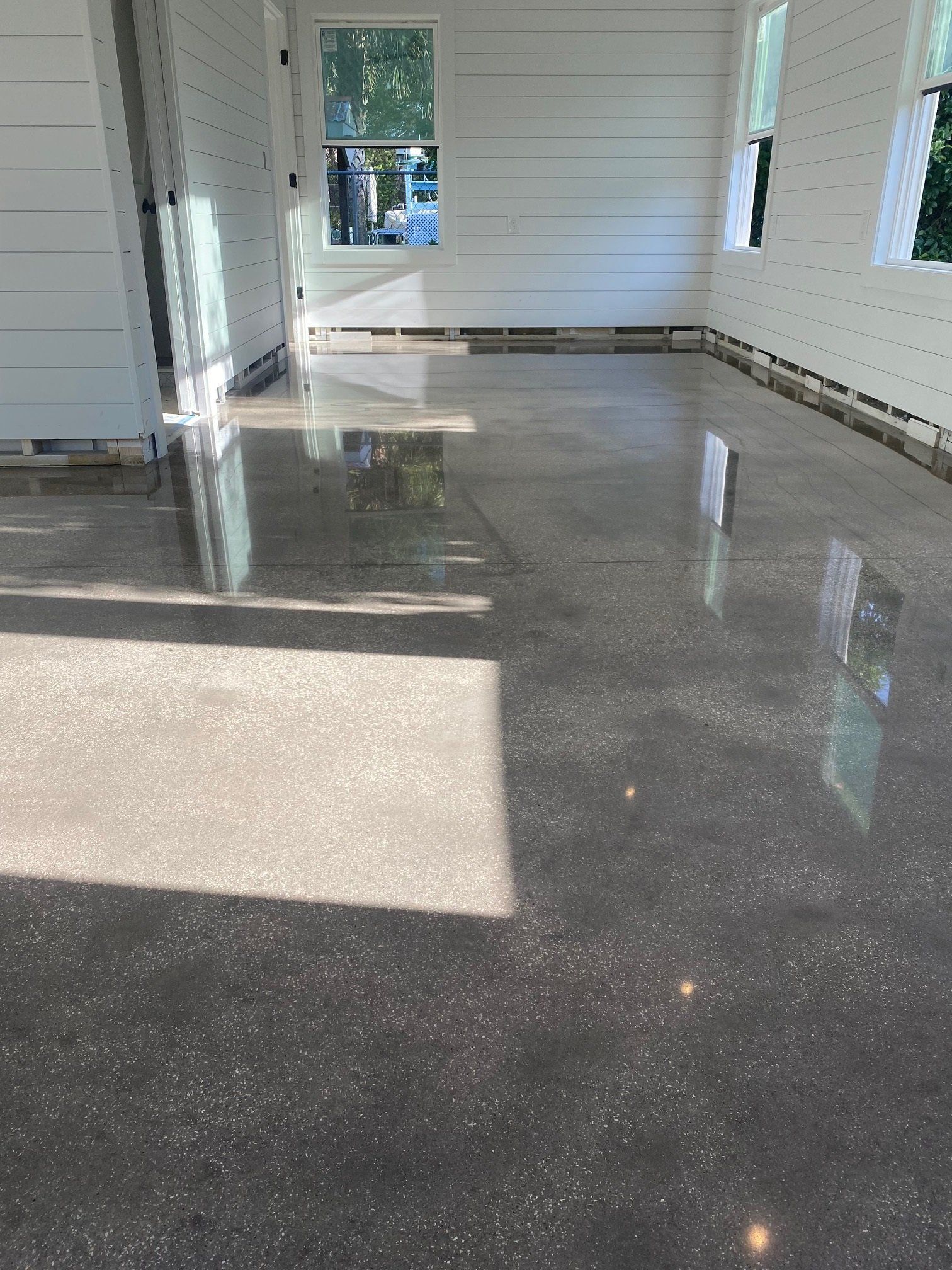 Polished concrete floor in a room with white shiplap walls and windows. Sunlight reflects off the glossy surface.
