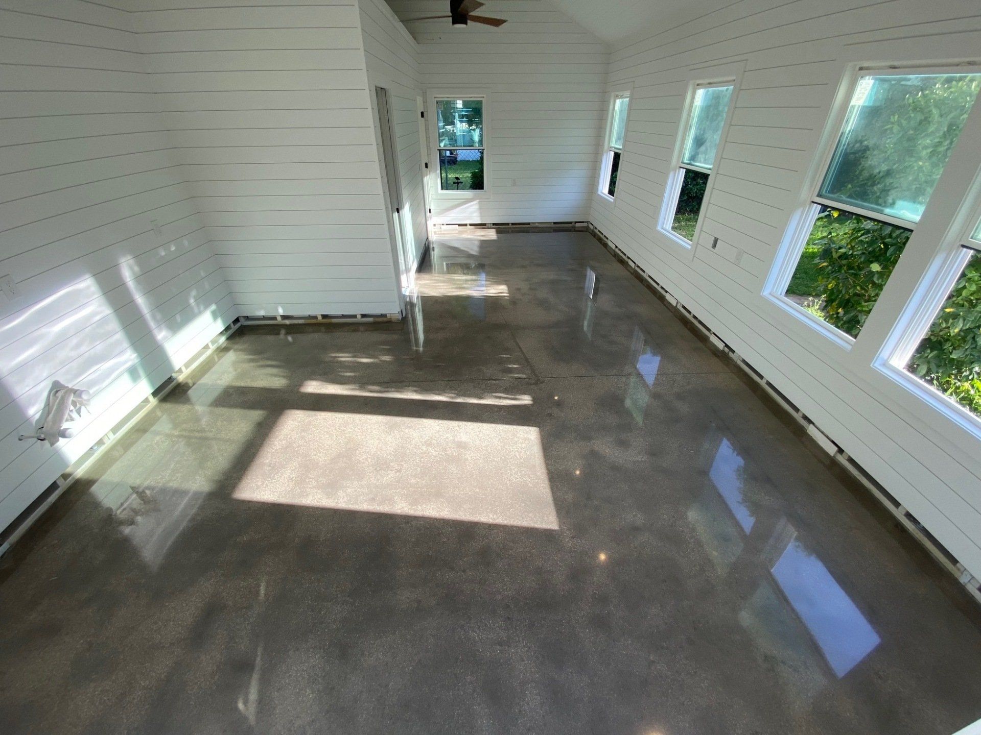 Sunroom with polished concrete floor, white horizontal shiplap walls, and bright windows overlooking greenery.