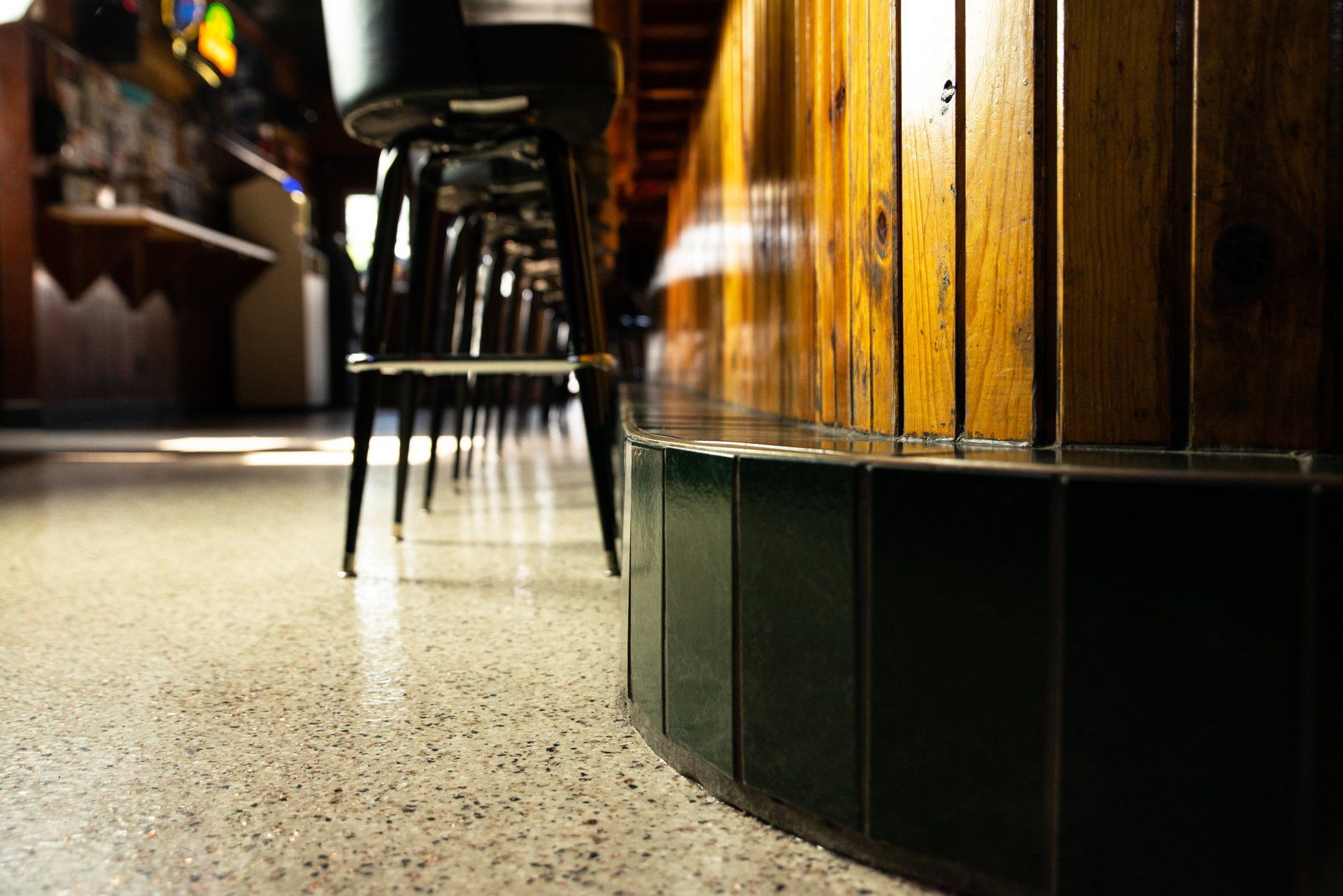 Low-angle view of a bar with a polished wooden counter and black bar stools. The floor is speckled.